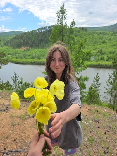 A young woman with light brown hair and blue eyes smiles at the camera while holding a bouquet of yellow flowers. She is wearing a grey t-shirt and purple shorts. In the background, a river flows through a lush green valley, with hills and trees covered in foliage. The sky is partly cloudy. The scene appears to be a scenic overlook. This location is likely in Atamanovka, Russia, known for its natural beauty and river landscapes.