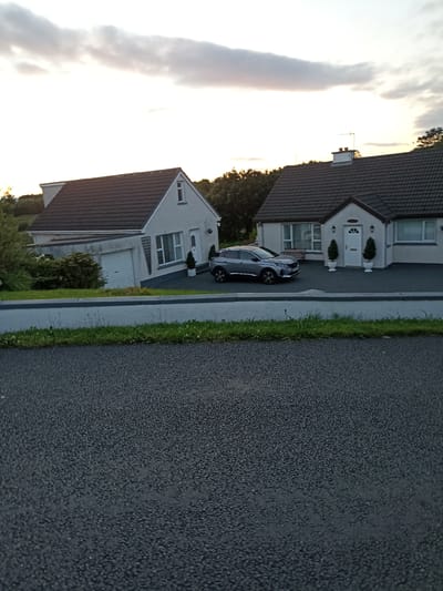 The image shows a street view of a residential area, likely in Harley Court, Ireland, during the evening. Two semi-detached houses with dark tiled roofs and light-colored exteriors are visible. A grey SUV is parked in the driveway of the house on the right. The house on the left has a garage with a white roller door. A concrete curb separates the road from a strip of green grass with some weeds. The road in the foreground has a dark asphalt surface. The sky above is partly cloudy with a mix of light and dark clouds, suggesting either dusk or dawn. Trees and foliage are visible behind the houses.