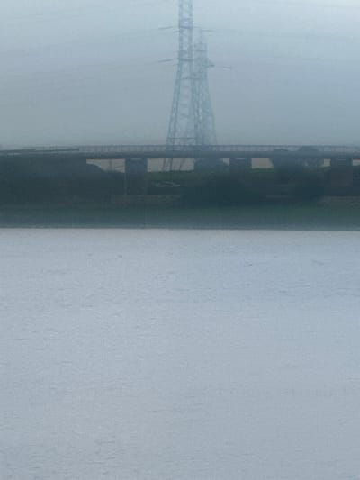 The image depicts a wide shot in Oumazza, Morocco, under hazy or overcast conditions. The lower half of the frame is dominated by a light gray, textured surface of a body of water, possibly a river or lake, showing subtle ripples. Above the water, spanning the mid-ground horizontally, is a dark, multi-span bridge with visible support pillars. Beneath the bridge, between the pillars, an area of dark vegetation is present, and a light-colored vehicle, possibly white, is partially visible. Further in the background, a tall, metallic lattice-structured electricity pylon stands prominently, rising against a uniformly light gray, obscured sky. No individuals or specific actions are discernible.