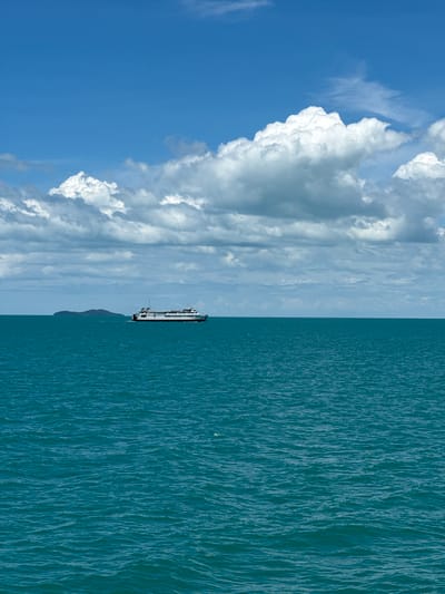 A ferry travels across a vast expanse of turquoise water under a bright blue sky dotted with fluffy white clouds. In the distance, a small, tree-covered island is visible on the horizon. The water is calm with gentle ripples, reflecting the sunlight. The ferry is a long, white vessel with multiple decks and windows, appearing to be traveling from left to right. The scene evokes a sense of tranquility and open space.