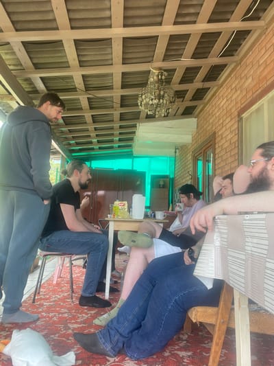 Six men are gathered on a covered outdoor veranda or porch in Vasilevka, Russia. The structure features a wooden slatted ceiling with a corrugated grey roof and an ornate, multi-tiered crystal chandelier hanging centrally. The right wall is constructed of red brick, containing a framed window or door. The floor is covered by a red and cream geometric patterned rug.

On the far left, a Caucasian male with short brown hair and a beard, wearing a dark grey hoodie and teal-grey sweatpants, stands with his hands in his pockets, looking down and to his right. Adjacent to him, a Caucasian male with long brown hair tied in a bun and a full beard sits on a chair, wearing a black t-shirt and blue jeans. He faces right, with his right arm bent and left arm resting on his lap.

At a small, light-colored wooden table in the center, a yellow bag of snacks (partially visible brand 'Хрусteam'), a small red container, a white plastic bucket, and several white mugs are present. A Caucasian male with dark hair, wearing a black baseball cap, a light purple t-shirt, and black shorts over white undergarments, sits at the table, drinking from a white mug. His right leg is crossed over his left, and he wears light green socks. Partially obscured behind the table, another male's legs are visible, clad in blue jeans and black socks.

To the right, a Caucasian male with short dark hair and light stubble, wearing a dark t-shirt, is reclined in a chair with his left arm behind his head. Further right, a Caucasian male with long dark hair, a full beard, and glasses is seated, wearing a dark t-shirt and blue jeans. His right arm rests on the striped armrest of his chair. A black object, possibly a mobile phone, rests near his arm.

A white power cable is visible running along the ceiling beams. A white plush object, possibly a goose-shaped pillow, rests on the rug in the bottom-left foreground. The scene is illuminated by natural daylight from the left, revealing greenery outside the structure. The men appear to be engaged in conversation and relaxation.