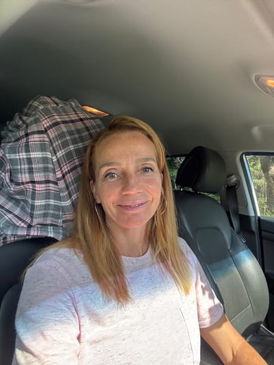In Portovoe, Russia, a woman with blonde hair and freckles, wearing a pink t-shirt, takes a selfie from the passenger seat of a car. Her hair is shoulder-length and slightly windswept, and she is wearing hoop earrings. In the background, a black car seat and a gray and pink plaid blanket are visible. Outside the car, trees can be seen through the window, suggesting a natural, possibly rural, setting. The lighting indicates it is daytime, with sunlight casting shadows and highlights on the woman's face and clothing.