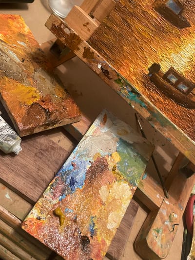 The image shows a close-up of an artist's workspace. In the upper left is a section of a textured painting on an easel, featuring warm hues and possibly depicting buildings or structures. Adjacent to the painting are several artist palettes splattered with various paint colors including white, yellow, brown, orange, blue, and green. A small paintbrush is resting near one palette. A tube of Winsor & Newton paint is partially visible in the bottom center. The background appears to be a wooden surface, possibly a table or part of the easel. The image appears to be taken indoors, under warm lighting. There is no specific information available in the image to identify that it is located in Maidstone, United Kingdom.