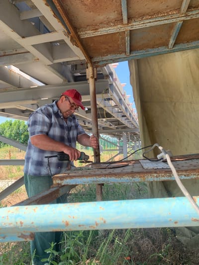 A man wearing a red cap and glasses is using an angle grinder to cut metal. He is wearing a plaid shirt and green pants. The scene appears to be outdoors under a metal structure. The background shows more metal structures, some greenery, and a clear blue sky. The metal structures show signs of rust. The man is focused on his work, and sparks are flying from the grinder. The image was taken in Pivdennoukrainsk, Ukraine.