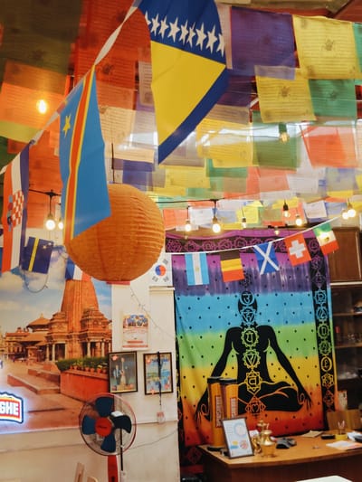The image appears to be an interior shot of a brightly decorated room, possibly a shop or a cafe, adorned with numerous flags and prayer flags hanging from the ceiling and walls. In the foreground, a red and blue electric fan stands on a stand. To the left, a large mural depicts a temple complex, likely in Nepal given the context of Kathmandu. The mural shows intricately carved reddish-orange structures with multiple levels and spires, set against a cloudy sky. Several potted plants are arranged in front of the mural.
The room is filled with various flags. Most prominent are the colorful prayer flags, draped in horizontal rows across the ceiling, featuring printed texts and symbols. Several national flags are also visible, including the flag of Bosnia and Herzegovina, the flag of the Democratic Republic of Congo, and the flag of Croatia. Smaller flags of countries like Argentina, Belgium, Scotland, Switzerland, and Mexico are also present, strung along a line.
A large, orange, spherical lantern hangs from the ceiling. On the wall to the right of the fan, there are three framed pictures. The top one is a poster for a "Cooking Class." Below it are two framed photographs, one showing a man and a dog, and the other showing people, possibly in a group.
Further to the right, a large, black tapestry hangs on the wall, featuring a stylized human figure with chakras depicted in rainbow colors. On a wooden desk in front of this tapestry, there are several objects including two cylindrical containers with labels, a small framed picture, a golden brass kettle or pot, and other desk supplies. The overall impression is one of a vibrant and culturally diverse space.