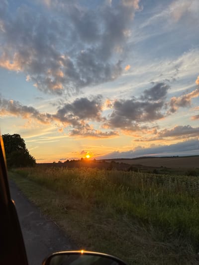 The image depicts a rural landscape at sunset, likely in or near Naraiv, Ukraine. The sun, a bright orange orb, is setting near the horizon, casting a warm glow across the sky and illuminating the clouds. The sky is filled with a mix of light and dark clouds, some tinged with pink and orange from the setting sun. In the foreground, a dark, unpaved road runs alongside a field of tall grass and vegetation. A large tree is visible on the left side of the image, silhouetted against the bright sky. In the distance, rolling hills and fields can be seen, with a few scattered trees and possibly buildings. The overall impression is one of natural beauty and tranquility at the end of the day.