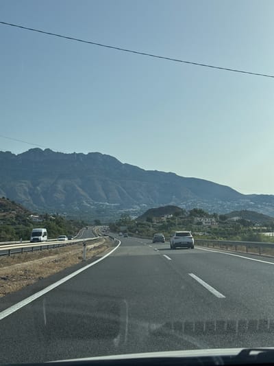 This media file captures a scenic view of a highway winding through a mountainous landscape, likely in Spain. The image is oriented vertically, with the highway occupying the right side of the frame and a large, rugged mountain range dominating the left. The sky is a pale, hazy blue, suggesting a clear but possibly warm day.
Several white vehicles, appearing to be vans or campervans, are traveling along the highway in the same direction. They are spaced out, indicating normal traffic flow. The highway itself is paved with asphalt and marked with white lines, including a broken white line on the right side of the lane and a solid white line bordering the shoulder. A metal guardrail runs along the edge of the highway.
The mountain is covered in sparse vegetation, with some areas appearing rocky and barren. Scattered houses and buildings are visible on the lower slopes of the mountain, suggesting a small village or community nestled into the hillside. The overall impression is one of a rural or semi-rural setting, with the highway providing a modern artery through the natural beauty of the terrain.
A thin, dark wire stretches diagonally across the upper left portion of the image, possibly a power line or telephone wire, adding a man-made element to the natural scenery. The lighting suggests it is daytime, likely late morning or early afternoon, given the bright but not harsh sunlight. There is no visible text in the image. The scene conveys a sense of travel and exploration, with the open road leading through an impressive natural backdrop.