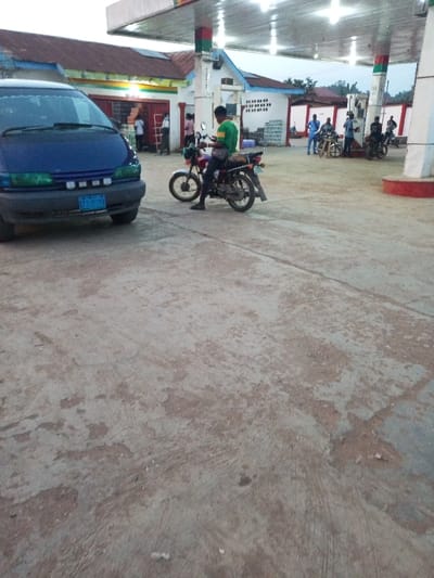This image captures an outdoor scene at a gas station in Aramoko-Ekiti, Nigeria, likely during late afternoon or early evening as artificial lights under the canopy are illuminated. The ground is a worn, light-colored concrete or asphalt.

In the foreground, a blue minivan is partially visible on the left, displaying a blue license plate that reads "KFS-771T". To its right, slightly further back, a man wearing a green and yellow striped shirt is seated on a red and silver motorcycle, appearing to be engrossed with a mobile phone.

The background reveals the main structures of the gas station. A large canopy covers several gas pumps where multiple individuals are gathered, many standing near their motorcycles. Behind the pumps, a single-story building with a reddish-brown corrugated roof and light-colored walls serves as a shop or office. Colorful horizontal stripes (green, yellow, red) adorn the building's facade, and a sign with unreadable text is visible below them. Stacks of crates, possibly containing beverages, are placed outside this building. The gas station pillars supporting the canopy are also painted with green, white, and red stripes. Several other people, predominantly men, are dispersed around the pumps and near the buildings, suggesting a busy period of activity such as refueling or socializing. The overall atmosphere appears casual and typical of a community gathering spot.