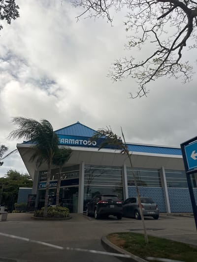 The image depicts the exterior of a Farmatodo pharmacy in the city of La Asuncion, Venezuela. The building features a modern architectural design with a prominent blue roof and a glass facade. Two cars, a black SUV and a gray hatchback, are parked in front of the pharmacy. Palm trees and other vegetation surround the building, and bare tree branches are visible in the sky, suggesting a cloudy or overcast day. A blue directional sign with a white arrow pointing left is present on the right side of the image. The scene appears to be a typical commercial area with parking facilities.