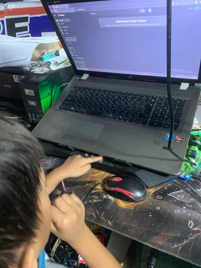 The image depicts an indoor scene, likely a home or office, with a young person interacting with a laptop. The laptop screen displays a YouTube interface, suggesting the person is browsing or watching videos. A black mouse with red accents is positioned on a patterned mousepad. The background includes a printer and stacks of paper, hinting at a cluttered workspace. The context of Sungai Meriam, Indonesia, is provided as the location. The overall impression is of a casual, everyday activity in a personal setting.