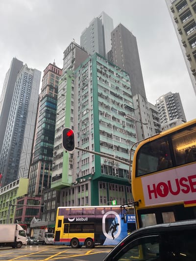 The image captures a street view in Hong Kong, China. Buildings of varying heights and architectural styles line the street, densely packed together. Many of the older buildings are characterized by numerous air conditioning units attached to their facades. A double-decker bus with advertising for "HOUSE" and "Webull" is prominently featured, along with various other vehicles on the street. A traffic light shows a red signal. The sky appears overcast, casting a diffused light over the scene. The composition of the image suggests the perspective of someone looking upwards from street level.