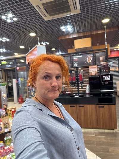 A selfie-style medium shot captures a woman with bright orange, curly hair and fair skin looking slightly upwards and to her right with a quizzical expression. She is wearing a light blue, collared, button-up shirt with a textured pattern and a black cord necklace. The background appears to be an interior retail space, possibly a convenience store or cafe, with a dark tiled ceiling with recessed lighting and air conditioning units. To her right, there is a coffee station with various dispensers and a black machine with a screen displaying an image. Shelving units stocked with products are visible behind the coffee station and to the left of the frame. The overall lighting is bright, indicative of an indoor commercial setting. There is no specific indication of the location being in the city of Можга, Russia, beyond the prompt.