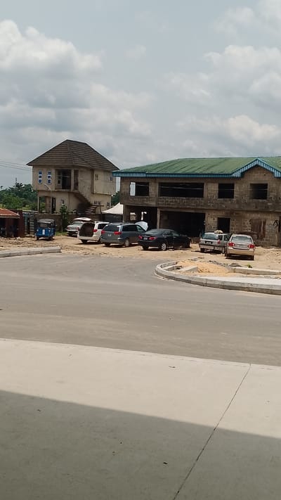 The scene depicts an urban area in Effurun, Nigeria. Two unfinished concrete buildings are visible in the background. The building on the left is a two-story structure with visible window openings but no glass. The building on the right is larger, also unfinished, with a green roof and open spaces where walls and windows would typically be. Several parked vehicles, including sedans and what appears to be a mini auto rickshaw, are in front of the buildings. The foreground consists of a newly constructed concrete road with a curved curb. The sky above is cloudy. Some people are visible near the building on the right.