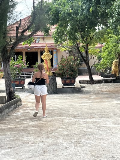 This image captures an outdoor scene at what appears to be a temple or cultural site, located in Cam Ranh, Vietnam. A woman, seen from behind, is the primary subject, walking on a light-colored, textured paved path. She is dressed in a black sleeveless top and white shorts, with light-colored shoes. Her hair is styled in a braid, and she is holding a white helmet in her left hand while also carrying a dark crossbody bag.
In the background, a building with a red-tiled roof is prominently featured. Its facade is richly decorated with intricate patterns in red, gold, and blue, and features several golden statues or relief sculptures, including a large, ornate figure near what appears to be an entrance. Dark windows or doorways are visible on the building's front.
The scene is framed by lush green trees with dense foliage, some of which appear tropical. Flowering bushes with pink blossoms add splashes of color near the building. On the paved path, a decorative black planter adorned with red and gold motifs is present. Further back, near another golden statue, a black bench can be seen with white text, which reads "BU TUC COC GIA." The overall setting suggests a daytime visit to a serene and culturally significant location.