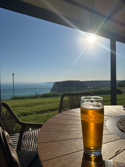 The image captures a serene scene outdoors, likely at a cafe or restaurant with a view. The primary subject is a glass of golden beer with a frothy head, positioned on a wooden table. The table surface has a distinct planked design. To the left of the table, a portion of a woven chair is visible, and a dark bag with a triangular logo rests on it.
The background reveals a breathtaking vista: a clear blue sky transitions into a hazy horizon overlooking a body of water, possibly the sea. Beyond the water, rolling green hills are visible, suggesting a rural or coastal setting. The sun is bright and high in the sky, casting lens flares and strong shadows, indicating it is daytime, likely late morning or early afternoon. The weather appears to be sunny and pleasant.
The scene evokes a sense of relaxation and enjoyment, perhaps someone taking a break to savor a drink while admiring the view. The focus on the beer and the scenic backdrop suggests a leisurely moment. The location context indicates this might be Kavarna, Bulgaria, known for its coastal beauty. There is no visible text in the image.