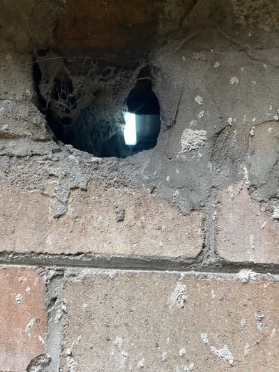 A close-up captures a hole in a weathered brick wall, revealing a bright light source within. Cobwebs fill the cavity, partially obscuring the view beyond. The bricks are heavily textured and show signs of age and wear, with crumbling edges and discoloration. Mortar lines are visible, and some bricks appear to be missing chunks. The light highlights the rough texture of the wall and the intricate web formations.