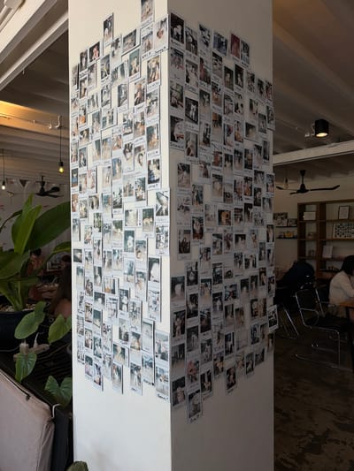 The image shows a white pillar adorned with a large display of polaroid photographs. The photos, predominantly featuring dogs, are arranged in a dense, overlapping collage. Many of the photos have handwritten names or captions written on their white borders. The pillar is located in an indoor setting, likely a cafe or restaurant, with exposed ceiling beams and lighting fixtures visible. In the background, patrons are seated at tables and chairs, and there is a shelf unit with books and other items. To the left, a large potted plant with broad green leaves is prominently displayed. The scene appears to be in a commercial establishment. The provided context identifies the location as Choeng Mon, Thailand.