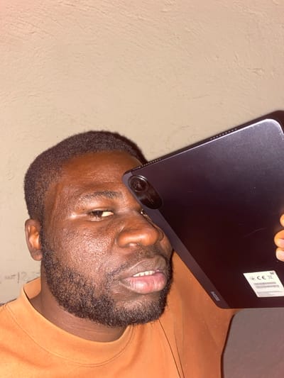 In the city of Udu, Nigeria, an adult male with dark skin, short dark hair, and a neatly trimmed beard is depicted. He wears an orange long-sleeved shirt. A dark gray Xiaomi Redmi tablet is held diagonally in front of his face, obscuring his left eye and upper forehead. The tablet's multi-lens rear camera module is visible near his right temple. The word "Redmi" and a white sticker with regulatory markings are discernable on the tablet's back surface. The man's right eye is partially visible, and his mouth is slightly open, revealing his lower teeth. The background consists of a plain, light-colored textured wall, illuminated by artificial light.