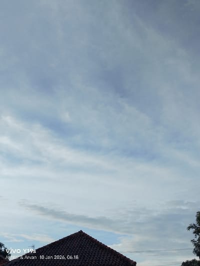 A low-angle shot presents a cloudy sky with streaks and patches of blue breaking through. A dark-red tiled roof occupies the lower center of the frame. To the lower right, a tree's leaves are partially visible. The sky dominates the composition, suggesting a wide-open space. The image has a soft, muted color palette. Text at the bottom left indicates the picture was taken with a vivo Y19s camera on January 10, 2026, at 06:18.