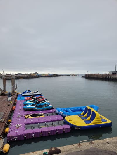 A fixed dock with wooden planks extends into the water. A modular floating dock made of purple plastic cubes is attached to the fixed dock and is covered with several jet skis. The jet skis are of various colors, including white, blue, red, and green. At the end of the floating dock, a large, inflatable water toy with blue and yellow sections is tethered. The water toy has seating areas. In the background, a harbor entrance is visible, with piers on either side. The sky is overcast with gray clouds. A person is visible on the fixed dock, dressed in casual attire. The location appears to be a waterfront area in Seogwipo-si, South Korea, likely a recreational or rental facility for watercraft.