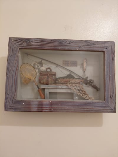 A rectangular shadow box with a rustic, painted wood frame hangs on a pale beige wall. The frame has a blue-grey wash with white speckles. Inside the box, arranged against a light-colored, possibly white or pale grey backdrop, is a collection of fishing-related items. These include a fishing rod, a fishing reel, a lure, a fish, and two nets. A small, woven creel and a photograph are also visible. The fishing rod is positioned diagonally, with the reel towards the top of the box. The fish stands upright with one net behind it, and one below. The presentation of these objects suggests a decorative or display purpose.
There is no information within the picture to suggest this image was taken in Cutler Bay, United States of America. This location has been given by request.