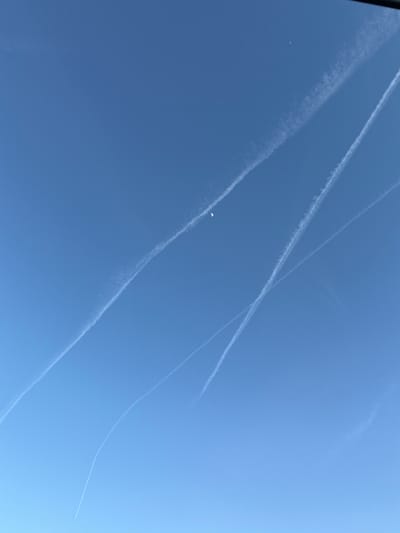 The image displays a clear daytime sky over Pétange, Luxembourg, dominated by multiple white contrails from aircraft. These long, linear vapor trails crisscross the blue expanse, with some appearing thin and sharply defined, while others are wider and more diffused, suggesting they have been present for varying lengths of time.
In the lower-left quadrant of the frame, a distinct, bright white crescent moon is visible against the blue sky. Further towards the middle-left, a tiny, bright white speck, possibly a distant aircraft or other airborne object, can be seen. The overall scene indicates clear weather conditions with no other clouds present, and the strong illumination points to the photograph being taken during daylight hours. No text is visible in the image, and no specific emotions are conveyed.