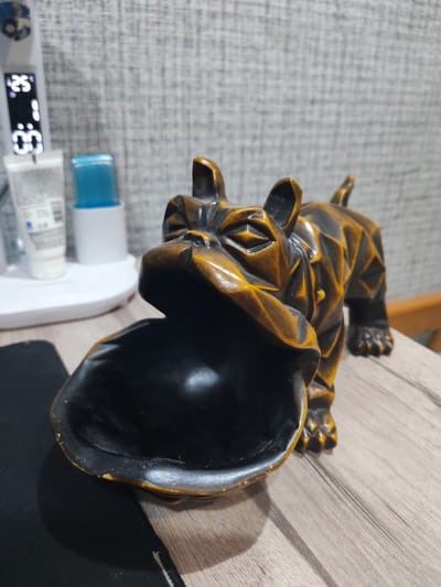 In Yalangach, Uzbekistan, a dog-shaped ashtray, with a geometric design and a dark, glossy interior, sits on a wooden surface. The ashtray has a weathered, metallic finish, with streaks of amber and black. Its mouth is open, revealing the dark interior. To the left, a white electronic device with a digital display showing "25°" and "1:00" is visible, along with two tubes of cosmetic products. The background features a textured gray wall.
