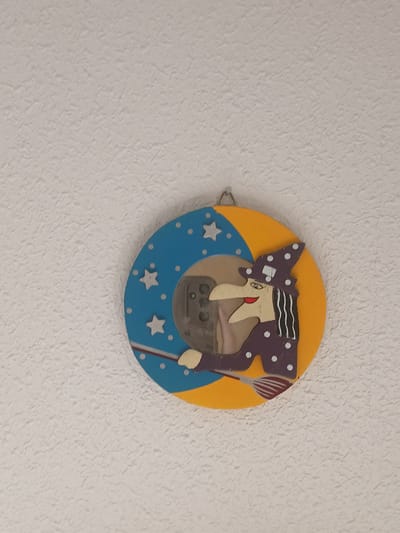 This media file features a round, decorative object resembling a clock or wall hanging, affixed to a textured, off-white wall. The object has a circular yellow background, overlaid with a blue crescent shape adorned with small white dots and several white stars. A cartoonish figure, seemingly a witch, is positioned in the center. The witch has a pointed nose, a red mouth, and eyes with long eyelashes. She wears a purple hat decorated with white polka dots and has black and white striped hair. In her hand, she holds a broomstick with red and white bristles. A metallic hook is attached to the left side of the object, suggesting it is meant to be hung. The overall design is whimsical and childlike, possibly a decoration for a child's room or a festive seasonal ornament. There is no visible text. The lighting suggests it is daytime.