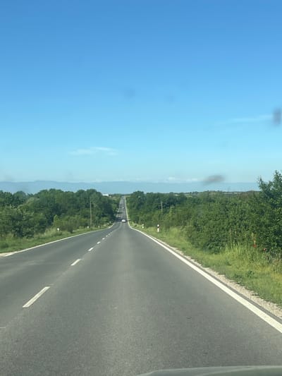The image depicts a straight stretch of asphalt road in Brezovo, Bulgaria, flanked by green vegetation on both sides. A clear blue sky occupies the left portion of the frame. In the distance, the road narrows to a vanishing point, with other vehicles visible along the asphalt. Road markings, including dashed white lines, are visible. The vegetation alongside the road appears to be a mix of trees and grasses. Mountains are faintly visible on the horizon. The image appears to have been taken from within a vehicle.