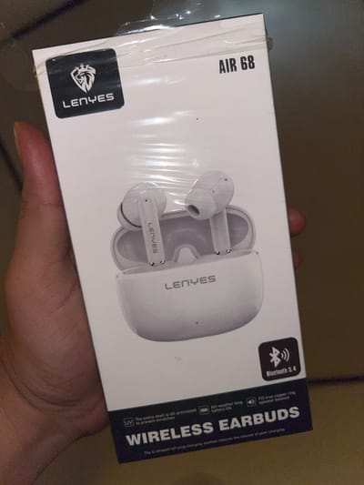 The image shows the front of a white box for "LENYES AIR 68 WIRELESS EARBUDS" held by a person's hand. The box is wrapped in clear plastic. A black sticker with a lion logo and "LENYES" is visible on the top left of the box. The product name "AIR 68" is printed in black on the top right. An image of white wireless earbuds and their charging case is printed on the front of the box. Several small icons and text descriptions of the product's features are also present, including "Bluetooth 5.4" and "UV The entire shell is UV-processed to prevent scratches". The background is dark and out of focus, suggesting an indoor, possibly dimly lit environment. There is no visible indication of the specific location within Keudematangkuli, Indonesia.