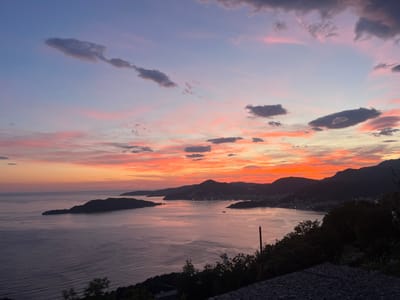 The image captures a sunset over a bay in Vojnići, Montenegro. The sky is filled with a mix of orange, pink, and purple hues. The foreground shows the bay with an island on the left. The dark silhouettes of mountains and hills are visible in the middle distance. Vegetation and a retaining wall are in the immediate foreground on the right.