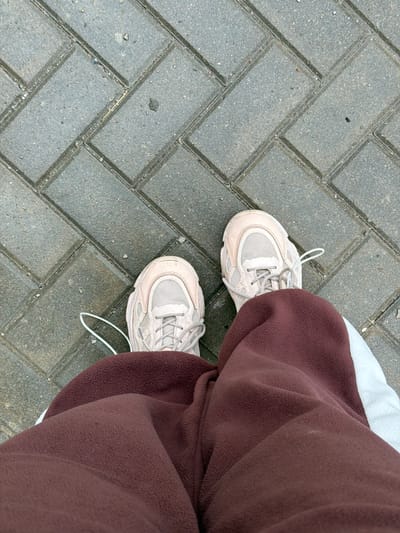 The image shows a first-person perspective of someone standing on a paved surface. The pavement consists of gray bricks laid in a herringbone pattern. The person is wearing light beige sneakers with white laces. The right foot is partially obscured by a pair of brown fleece pants. A small section of white fabric is visible near the top of the frame, likely part of the clothing. The background is composed of the textured gray pavement. The location cannot be determined from the image.