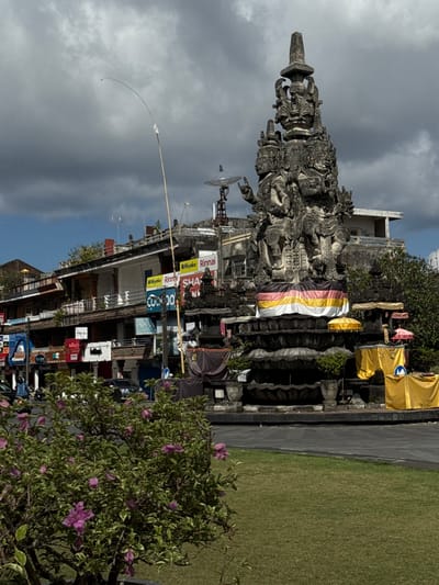 In Klungkung, Indonesia, a large, intricately carved stone monument stands prominently in a city square. The monument, adorned with multiple figures and decorative elements, is draped with a colorful fabric featuring stripes of white, yellow, and pink. Several smaller, ornate structures and potted plants surround the base of the monument, some covered with yellow fabric. A tall, slender bamboo pole with a white streamer extends upwards from behind the monument into the cloudy sky. Across from the monument, a row of multi-story buildings with balconies and awnings lines a street. Various signs, including "Miyako Rinnai" and "SHAI," are visible on the building facades. A paved road is in the foreground, with a patch of well-maintained green grass. A few vehicles, including a black car and a motorcycle with a rider, are present on the road. In the lower left corner, a bush with green leaves and delicate purple flowers is in bloom. The overall scene depicts a public space with a significant cultural monument, commercial establishments, and everyday urban activity under a dramatic sky.
