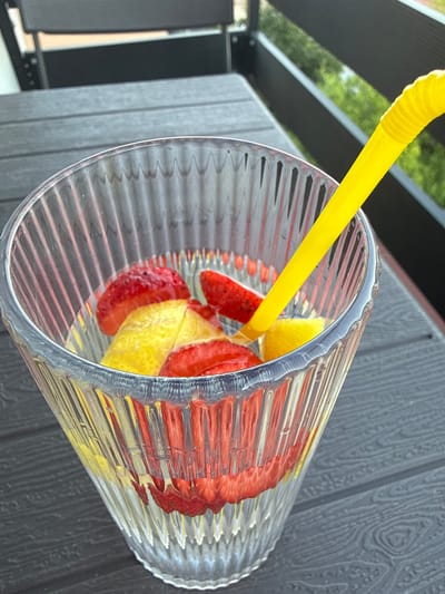 A close-up shot shows a clear, ribbed glass tumbler filled with sliced strawberries and lemon wedges. A bright yellow plastic straw is inserted into the drink, angled towards the top. The tumbler is tilted on its side, resting on a dark, textured surface that appears to be outdoor furniture, possibly a table. In the background, blurry elements suggest an outdoor setting with greenery and possibly some black structures. The lighting suggests it is daytime, with natural light illuminating the drink and its contents. The overall impression is that of a refreshing beverage prepared for outdoor enjoyment. There is no visible text in the image. The location context provided is Sandanski, Bulgaria.