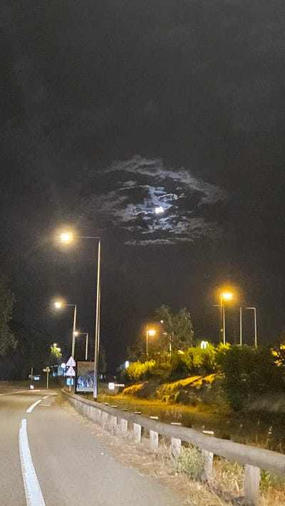 The image depicts a nighttime scene on a road likely near Privas, France, illuminated by streetlights and the moon. The road, with a white dividing line, curves to the left in the foreground. A metal guardrail runs along the right side of the road. Beyond the guardrail, a vegetated embankment is visible, with some areas illuminated by artificial lights. Several streetlights are present, casting a yellow glow. In the upper portion of the image, the moon is partially obscured by clouds, creating a dramatic effect. Signs are visible along the roadside, including pedestrian crossing signs and what appears to be a McDonald's sign in the distance. No people are visible in the frame. The overall impression is of a quiet, somewhat eerie, roadside environment at night.