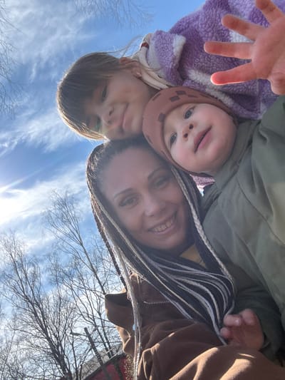 A selfie shows a woman with two children outdoors against a backdrop of a blue sky with wispy clouds and bare tree branches. The photograph is taken from a low angle, looking upwards. The woman is smiling, has light skin, and braided hair. One child wears a pink knit cap and a purple, textured jacket and extends their hand towards the camera. The other child has shoulder-length brown hair. The location is inferred to be Yaroslavl', Russia, based on the prompt, but there are no explicit indicators of the city visible in the frame.