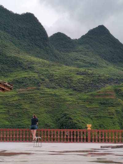 A person stands on a concrete balcony overlooking a verdant, terraced mountainside in Lũng Cú Commune, Vietnam. The person, viewed from behind, wears a dark t-shirt and denim shorts, leaning on a railing. A small, metal stool is positioned near them on the wet balcony floor, which reflects puddles of water. The railing is ornate and red-brown in color. Behind the railing, the mountains rise steeply, covered in dense green vegetation, with visible terraces carved into the slopes. The sky is overcast and gray. A portion of a building's tiled roof is visible on the left side of the frame.