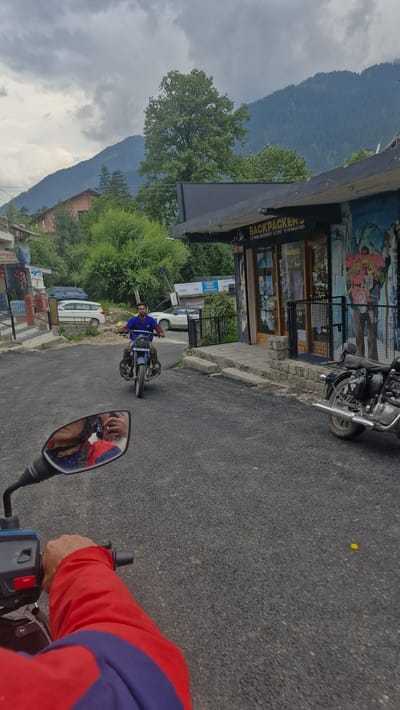 A person is riding a motorcycle on a paved road in Manali, India. The image is taken from the perspective of another motorcycle rider behind the first one, showing their red and blue jacket and the handlebars of their bike. The first rider is wearing a blue shirt and is on a blue motorcycle. They are riding past a storefront with a mural on its facade and a sign that reads "BACKPACKER'S TREKKING EQUIPMENT STORE". Mountains are visible in the background, partially obscured by trees and clouds, suggesting a mountainous, possibly cloudy day. The scene appears to be a street in a town or village, with other vehicles and buildings present. The overall impression is of a casual journey through a scenic location.