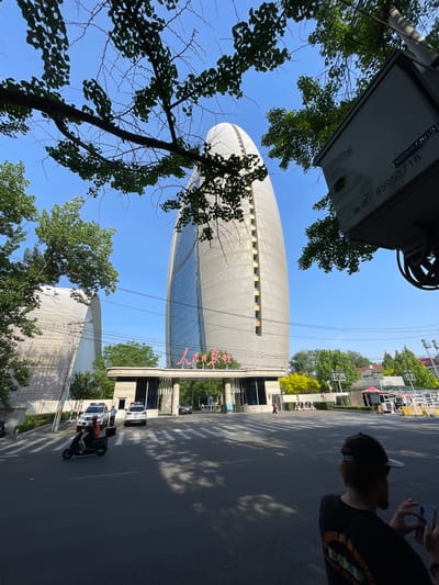 The image, taken in Chaoyang District, China, shows an urban scene with a prominent building dominating the view. The building is tall, elliptical, and light-colored, with rows of windows and Chinese characters on its facade. A road runs parallel to the building, with a pedestrian crossing visible. The sky is clear and blue, and the sun casts shadows across the scene. A person with a cap on is seen on the right of the scene. Some trees in full foliage are present in the image, partially obscuring the building and adding depth to the scene. A vehicle resembling a motor scooter with a rider is seen on the road. An overhead sign is partially visible at the top of the frame.
