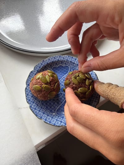 In Chang Phueak, Thailand, on a white countertop, two small, round, dark brown, seed-covered food items sit on a blue and white patterned plate. The food items appear to be made from a ground mixture, possibly meat or a plant-based alternative, with large, flat, green seeds pressed into their surfaces. A person's hands are interacting with the food. One hand, with a finger bandaged, is holding one of the food items, while another hand, with fingers extended, is delicately pressing seeds onto the other food item. In the background, a large, light gray, speckled plate is partially visible. The lighting suggests an indoor environment.