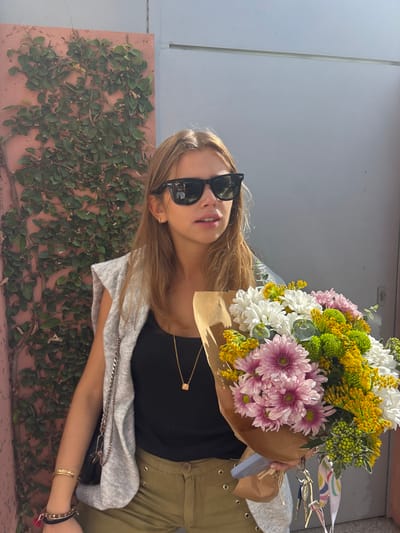 A young adult female is depicted standing outdoors in what appears to be a brightly lit urban or suburban environment, within the city of Aldoar, Foz do Douro e Nevogilde, Portugal. She has long, light brown hair and is wearing black Wayfarer-style sunglasses, a black ribbed tank top, a light grey sleeveless knit vest, and olive green trousers featuring decorative metal eyelets along the sides. A gold chain necklace with a small, light-colored cubic pendant hangs around her neck, and her left wrist displays multiple bracelets, including a wide gold-colored cuff and beaded bands. She is holding a large, mixed bouquet of white, pink, yellow, and green flowers, primarily chrysanthemums, wrapped in brown kraft paper. A set of keys with a colorful patterned ribbon is attached to the flower wrapping. The background to her left features a textured, reddish-pink wall densely covered in dark green ivy, while the background to her right consists of a plain, light grey wall or metal panel.