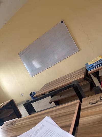 The image depicts an indoor scene, possibly a classroom or office, in the city of Baganakwo, Nigeria. The walls are painted a pale yellow color. A whiteboard is mounted on the wall, with some writing faintly visible. Several desks are arranged in a row, with black metal frames and light brown wooden tops. A stack of blue books is visible on one of the desks. A sheet of paper with printed text is partially visible on the right side of the frame. There are no people visible in the scene.