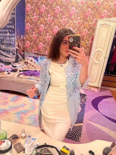 A young woman with long, dark hair is taking a mirror selfie in a bedroom. She is wearing a light-colored, form-fitting dress and a light blue cardigan adorned with rhinestones. She has a silver necklace with a blue pendant. Her nails are painted with gold glitter polish.

The room is decorated with floral wallpaper. A large wardrobe with ornate details stands to the right. A bed with unmade bedding and pillows is visible in the background. A fluffy, multicolored rug is on the floor in front of the bed. A desk or vanity table in the foreground is cluttered with various cosmetic items, including creams, tubes, a watch, and a makeup compact. A smartphone with a decorative case is being used to take the picture. The lighting suggests an indoor setting, possibly during the daytime. The location is described as Krasnoe-na-Volge, Russia, though no visual cues in the image directly support this specific location.