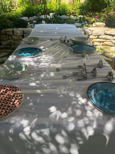 An outdoor dining table is set in a garden in Kumbor, Montenegro. The table is covered with a white tablecloth and features place settings with plates, bowls, and candles. Several white candles are placed in clear glass and silver candle holders, arranged in rows. There are also blue and red-patterned decorative bowls and plates on the table. The garden setting is visible in the background, with stone walls, lush green plants, and sunlight dappling through the leaves, creating shadows on the tablecloth.