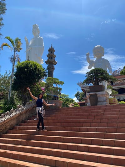 The image shows a young woman standing on a set of terracotta stairs with her arms outstretched. She is wearing a dark blue tank top, black leggings, and white sneakers. A light-colored jacket is draped over her shoulders. She is looking towards the camera and appears to be smiling, conveying a sense of joy and freedom.
In the background, two large white statues are visible against a clear blue sky with scattered white clouds. The statue on the left is a seated figure with a serene expression, wearing traditional Asian attire and a headdress. The statue on the right appears to be a smaller, childlike figure in a dynamic pose. A tall, multi-tiered pagoda-like structure is also visible between the statues. Lush green trees and flowering bushes surround the statues, adding to the vibrant atmosphere.
The scene is set outdoors, likely during the daytime given the bright sunlight and clear sky. The terracotta stairs suggest a temple or religious site. The overall impression is one of cultural tourism and enjoyment of a scenic and spiritual location in Thôn 5, Vietnam.