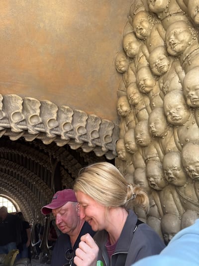 Two individuals are photographed within the HR Giger Bar in Epagny, Switzerland. A woman with blonde hair is in the foreground, her head tilted toward the right. A man wearing a maroon baseball cap is behind her, partially obscured. The architecture of the bar is prominently featured, characterized by skeletal structures and multiple sculpted faces on the ceiling and walls. Additional patrons are visible in the background, partially out of focus.