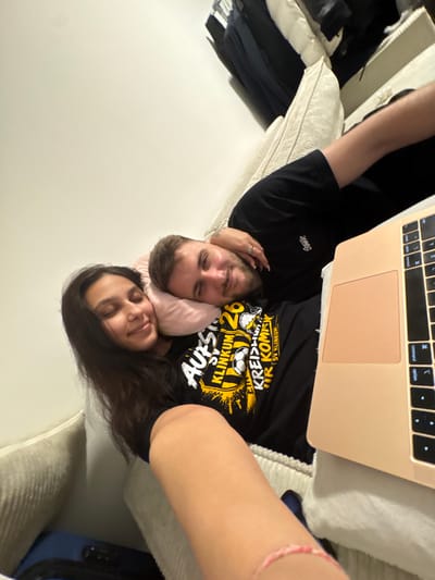 This image depicts a couple lounging on a sofa, with a laptop positioned in front of them. The woman is lying down, her head resting on a pink pillow, her eyes closed. The man is lying next to her, with his head also on the pillow, looking towards the camera. They are both wearing dark t-shirts. The t-shirt worn by the man has a graphic design with text that appears to be in German. The sofa is upholstered in a light-colored, ribbed fabric. In the background, on the left, there are clothes hanging on what appears to be a closet rod. The scene is indoors, likely in a residential setting. The presence of the German text on the t-shirt strongly suggests the location is in Germany. Mönchengladbach is a city in Germany, making it a plausible location for this scene.