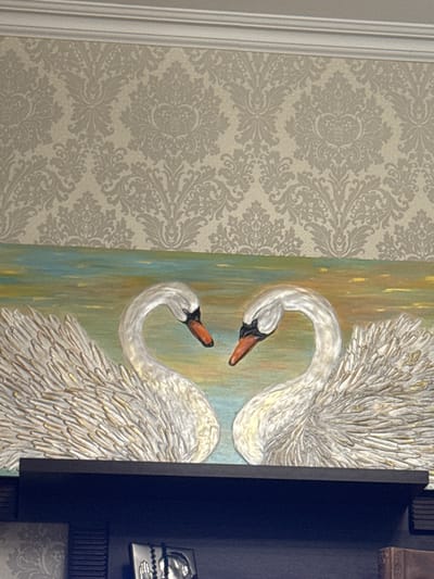 The image depicts a close-up of a textured painting featuring two swans. The swans are predominantly white with gold accents, rendered with thick impasto strokes that give them a three-dimensional appearance. The background of the painting consists of vertical streaks of color, primarily in shades of blue, green, and yellow, suggesting water or a stylized landscape. The painting is framed on the right by a dark, flat surface, possibly a cabinet or a wall panel, and on the left by a section of damask wallpaper. The wallpaper is a light beige color with a subtle, repeating pattern in a slightly darker shade of beige. To the far left, a portion of white molding or trim is visible, running vertically. The overall aesthetic is one of decorative art within an interior setting. There is no discernible activity or event taking place, as the image focuses on the artwork and its immediate surroundings. No text is visible within the frame. The lighting appears to be indoor, with soft illumination that highlights the texture of the painting. The location context of Slonim, Belarus, is provided but not visually evident in the image itself.