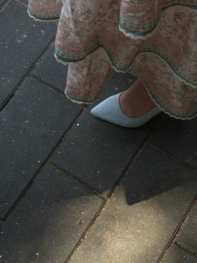 The image shows a close-up, overhead view of a person's foot wearing a light blue, pointed-toe heel. The shoe is positioned on a dark grey tiled surface, likely an outdoor walkway or patio. The person is also wearing a flowing skirt with a paisley pattern in shades of pink, light blue, and cream, with a decorative trim. The fabric of the skirt is draped and ruffles around the shoe.
Sunlight casts a shadow on the tiled ground to the right of the frame, suggesting it is daytime. The tiles are rectangular and laid in a pattern. Some small debris and lighter colored specks are visible on the surface of the tiles.
The scene is static, with no clear activity depicted beyond standing. The focus is on the texture and detail of the clothing and footwear against the background of the ground. The lighting and composition give a sense of casual elegance. The location context of Frankfurt, Germany, does not provide specific visual cues within this close-up shot. There is no visible text in the image.