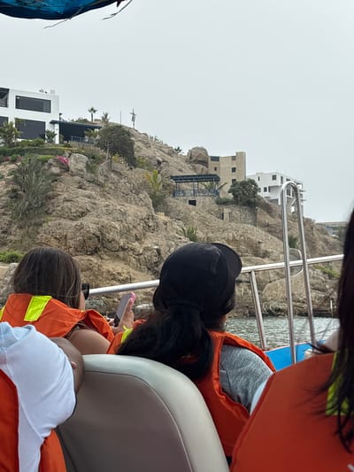 People are on a boat in Ancón, Peru, near a rocky coastline with buildings. The passengers are wearing life vests. One person is using a smartphone. A baby is lying down on a seat. The day appears overcast.